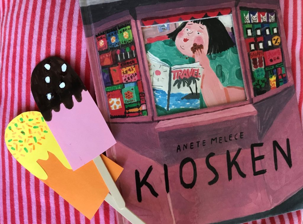 Bilderboken Kiosken av Anete Melece ligger på ett rosarandigt tyg. Bredvid ligger två pinnglassar av papper.