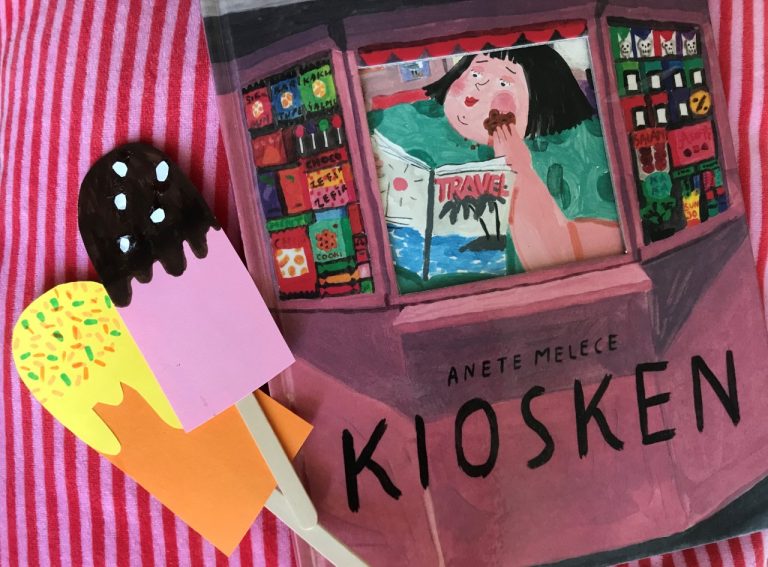 Bilderboken Kiosken av Anete Melece ligger på ett rosarandigt tyg. Bredvid ligger två pinnglassar av papper.