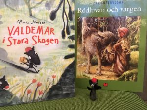 Framsidan på två böcker: Valdemar i stora skogen och Rödluvan och vargen. Framför böckerna en liten djurfigur med röd mössa.